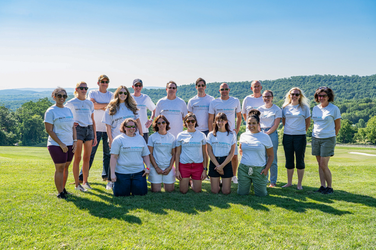 Group photo of KAA management members at corporate retreat, outdoors with green hills in the background, wearing branded white T-shirts.